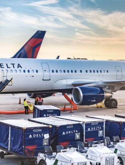 A Delta aircraft on the tarmac at sunset, ground crew loading cargo containers beside it.