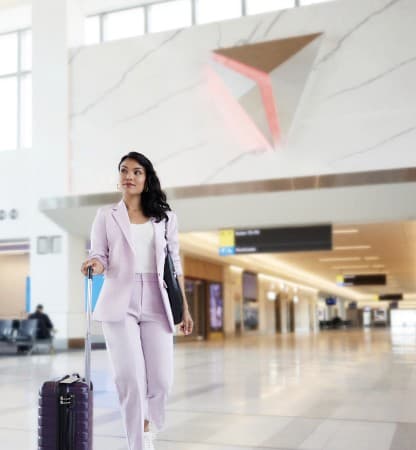 A woman wheeling a suitcase through a terminal past a large triangular Delta wordmark graphic on the wall.