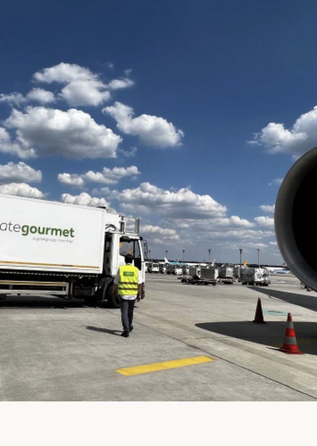 A Gate Gourmet high-loader truck on an airport apron, an engineer walking towards it past a parked aircraft's engine.
