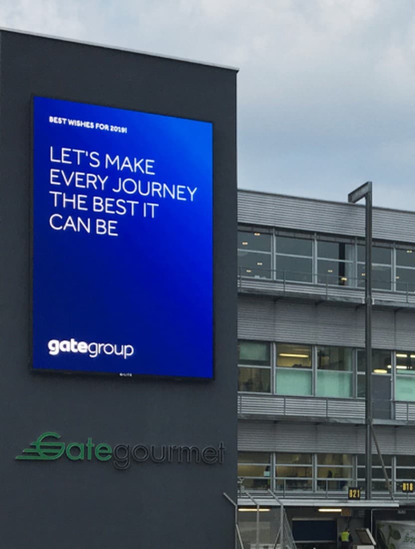 A gategroup-branded LED screen mounted on an office building facade, with Gate Gourmet signage below it.