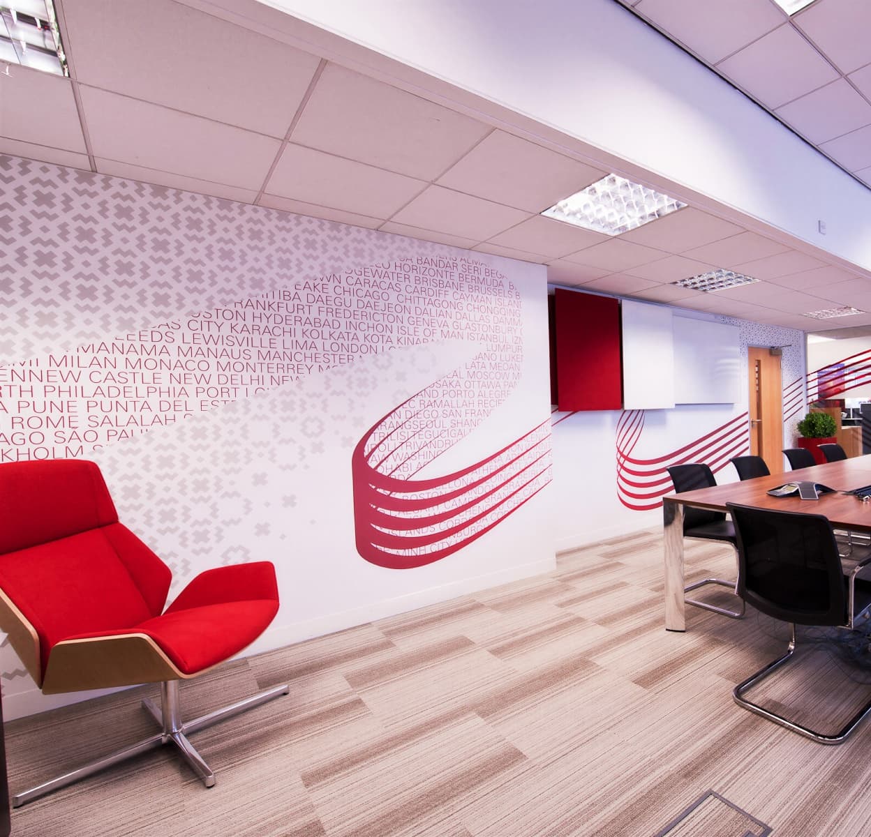 A meeting room with a red lounge chair against a wall printed with a red ribbon graphic and a field of city names.