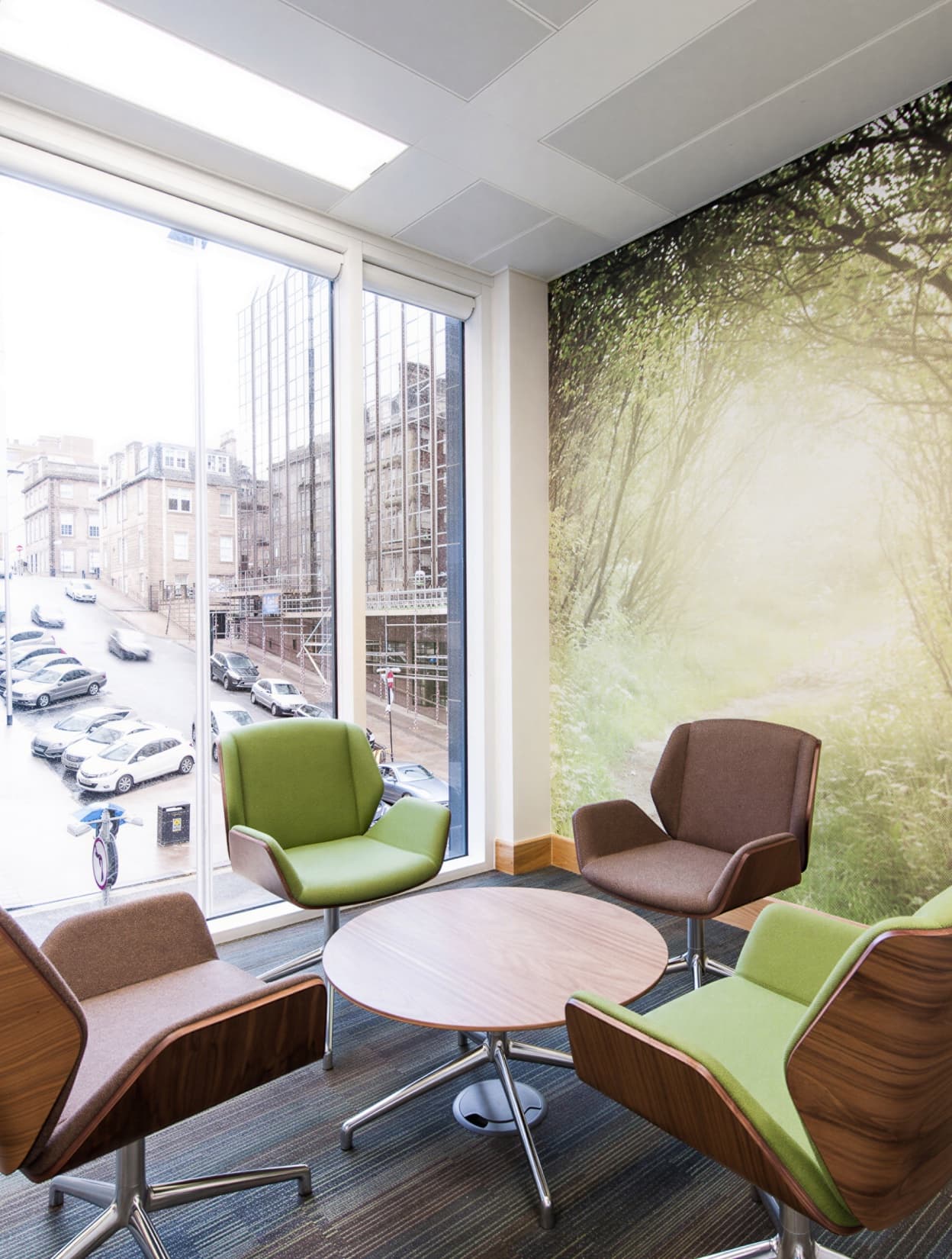 A window lounge with green and brown chairs around a low table, a forest mural covering the far wall.