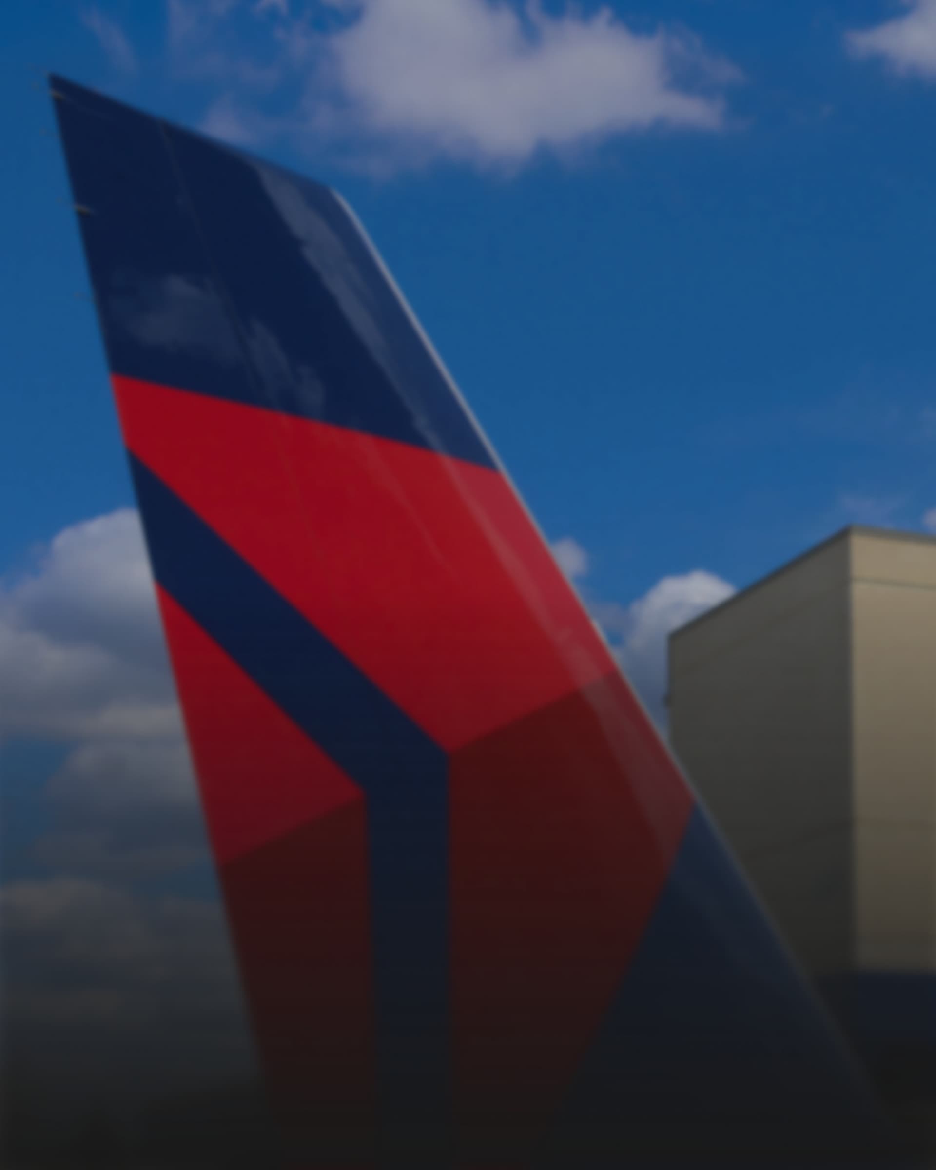 The tail fin of a Delta aircraft against a blue sky, an airport building visible behind it.