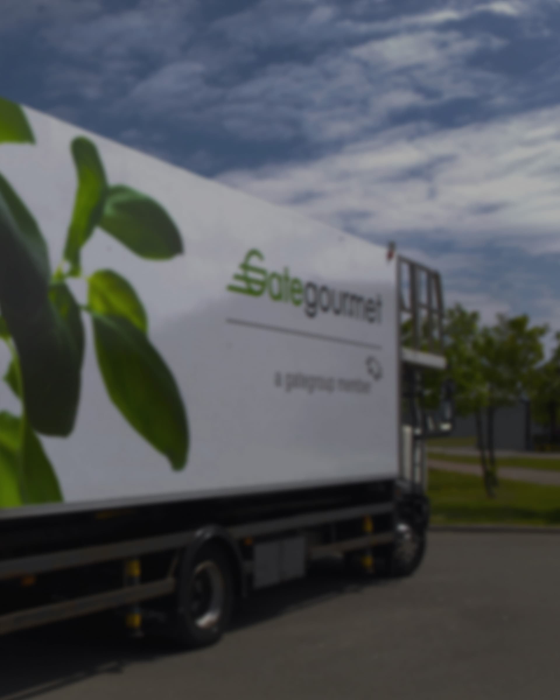A Gate Gourmet high-loader truck with the leaf livery on its side, driving under a blue sky with scattered clouds.
