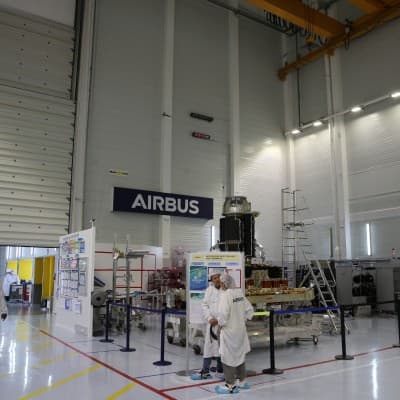 An Airbus assembly hall, the wordmark on the wall behind staff in white coveralls working on the floor.