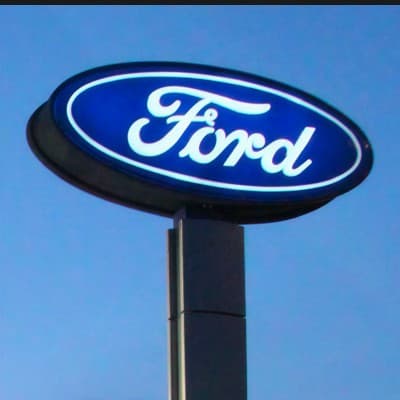 An illuminated Ford oval sign on a tall pole against a blue sky.