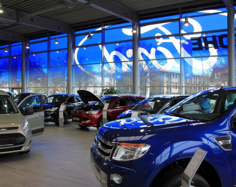 Inside a Ford showroom at dusk, cars on the floor and the Ford wordmark visible through the glazing behind them.