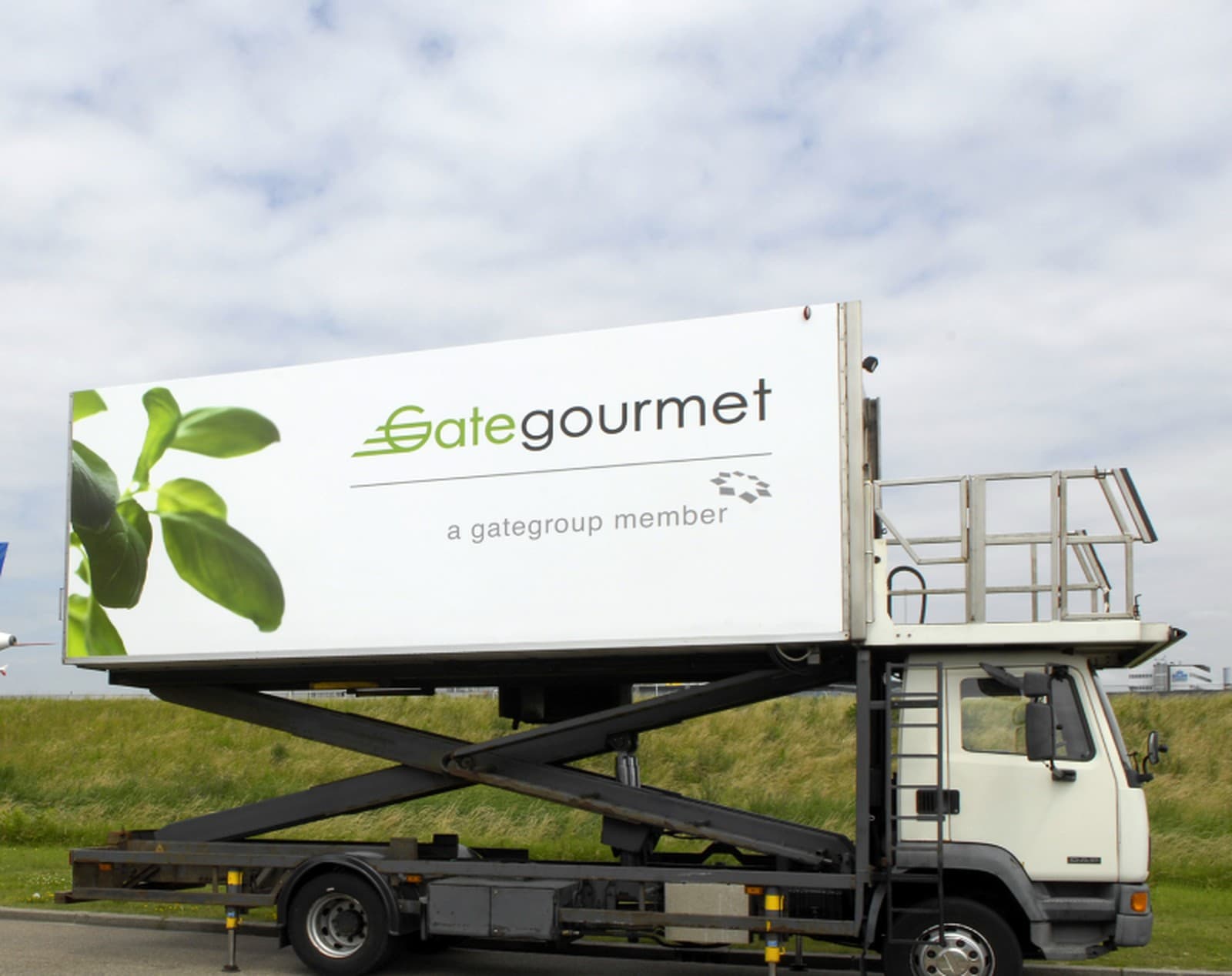 A Gate Gourmet high-loader raised on its scissor lift, the leaf livery visible on the container body, parked on grass beside an airfield.