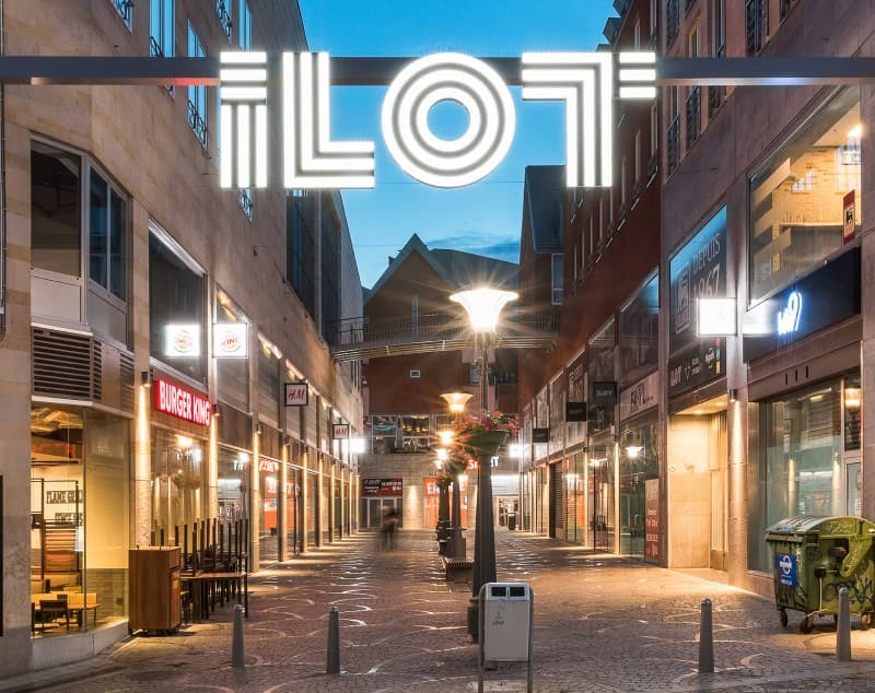 An illuminated ILOT sign spanning a street in the evening, shopfronts lit below.