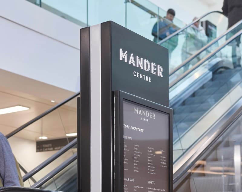 A Mander Centre directory totem beside the escalators, shoppers descending behind it.