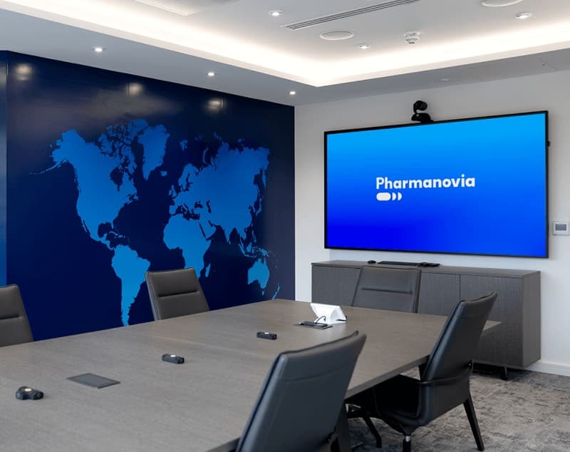 A meeting room with a world map graphic on one wall and a screen displaying the Pharmanovia wordmark.