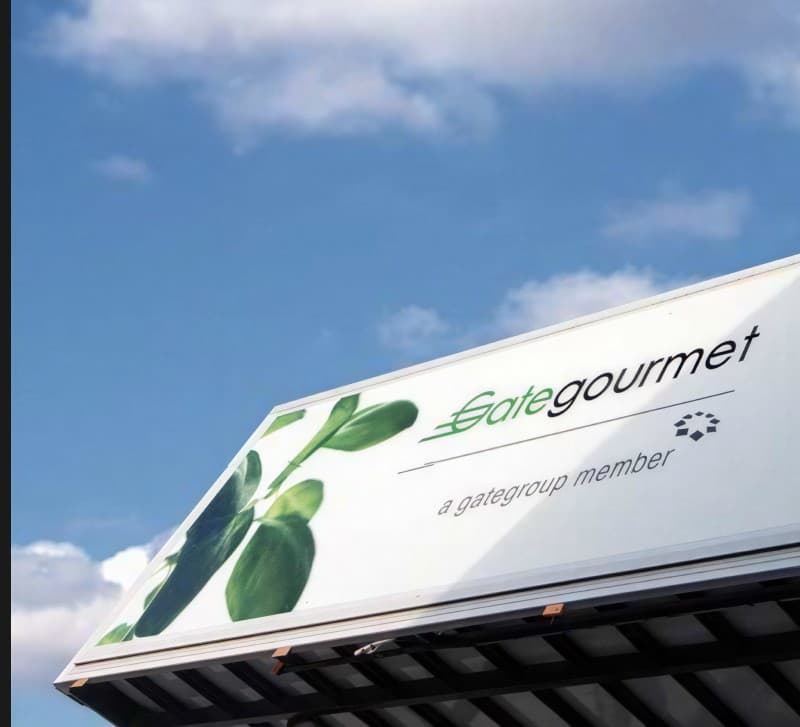 A GateGourmet high-loader truck showing the leaf livery and wordmark on its side, parked under a blue sky.
