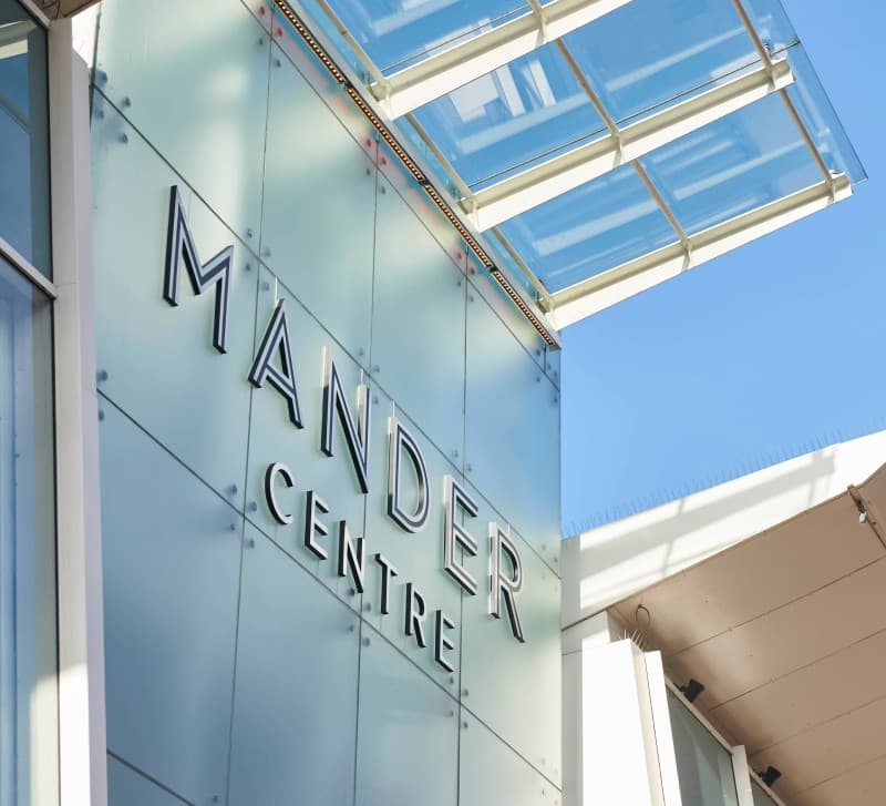 The Mander Centre entrance facade in daylight, its name lit above the canopy against a blue sky.