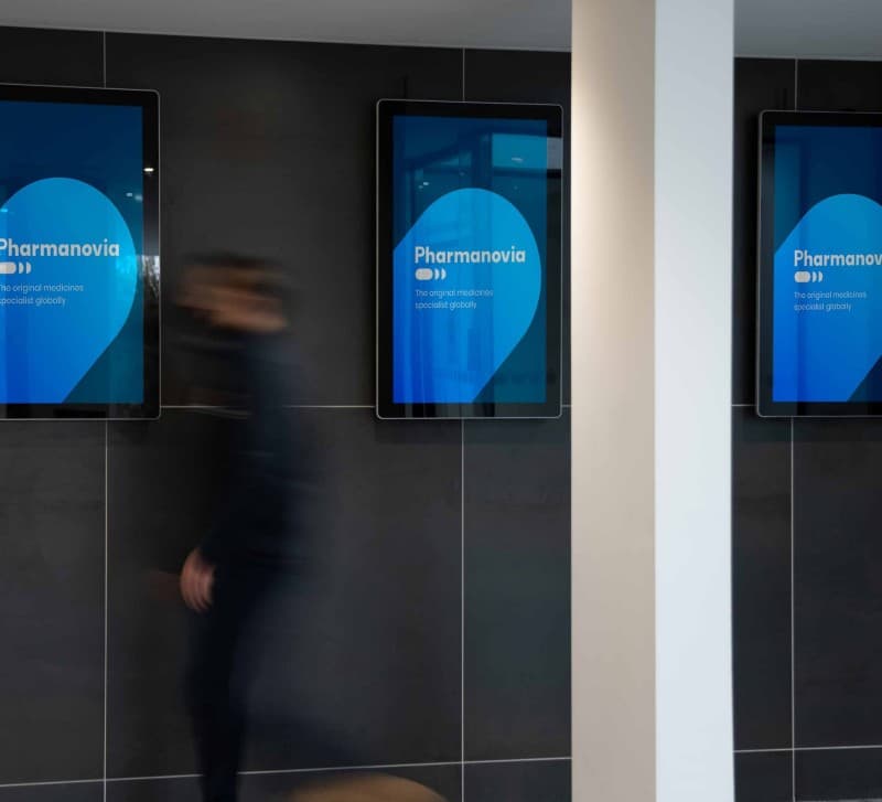 Three wall-mounted digital screens in a corridor, each showing the Pharmanovia wordmark.