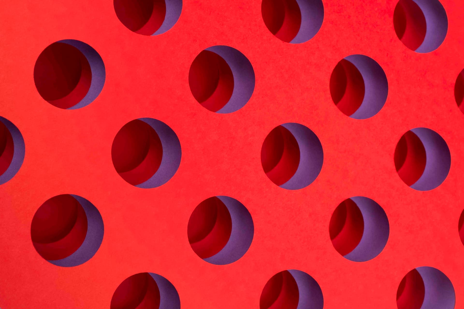 Repeating pattern of circular openings cut into a red surface, casting purple shadows