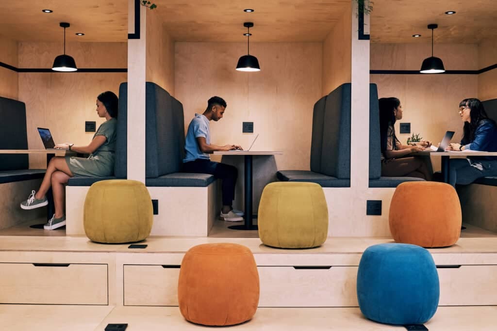 Office booths in pale plywood with people working at laptops, coloured stools in the foreground